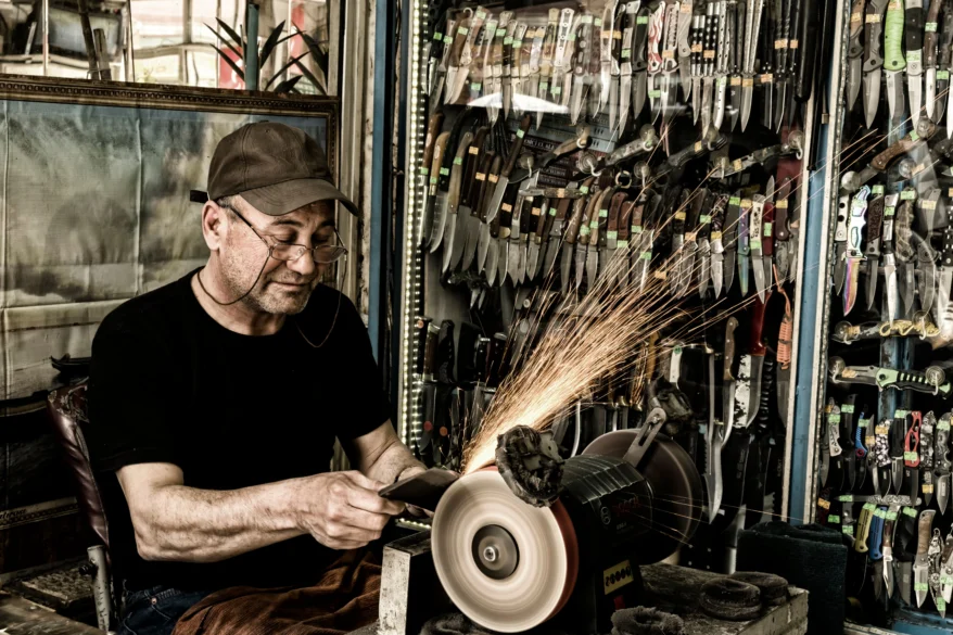 Knife maker sharpening a blade on a grinding wheel inside a workshop lined with knives.