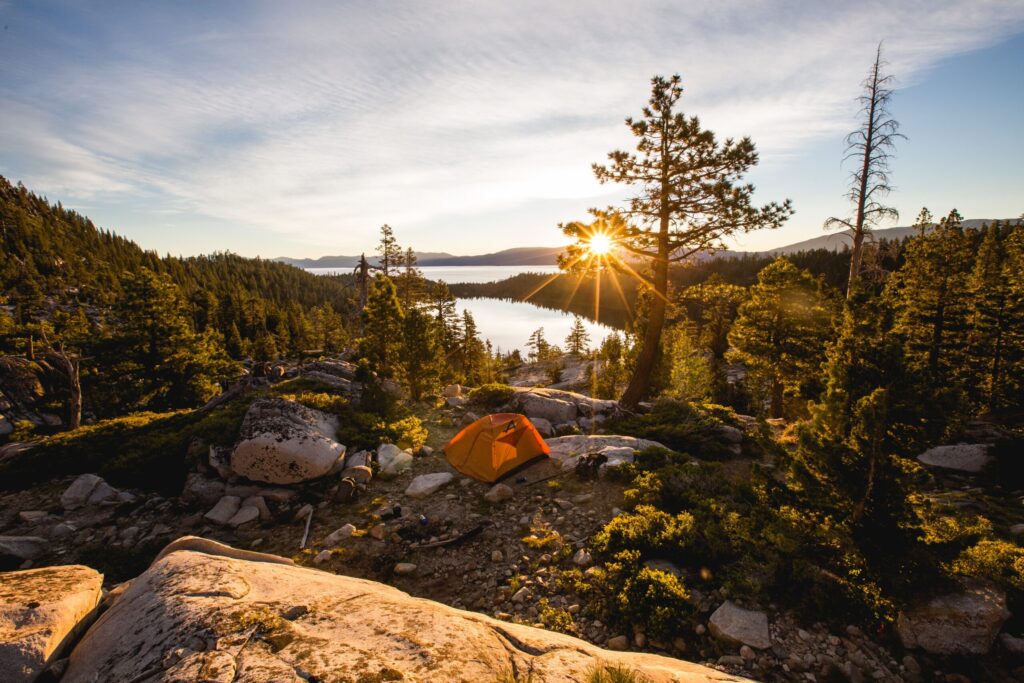 Golden sunset over a pine forest campsite beside a mountain lake, with an orange tent nestled among rocks and trees.
