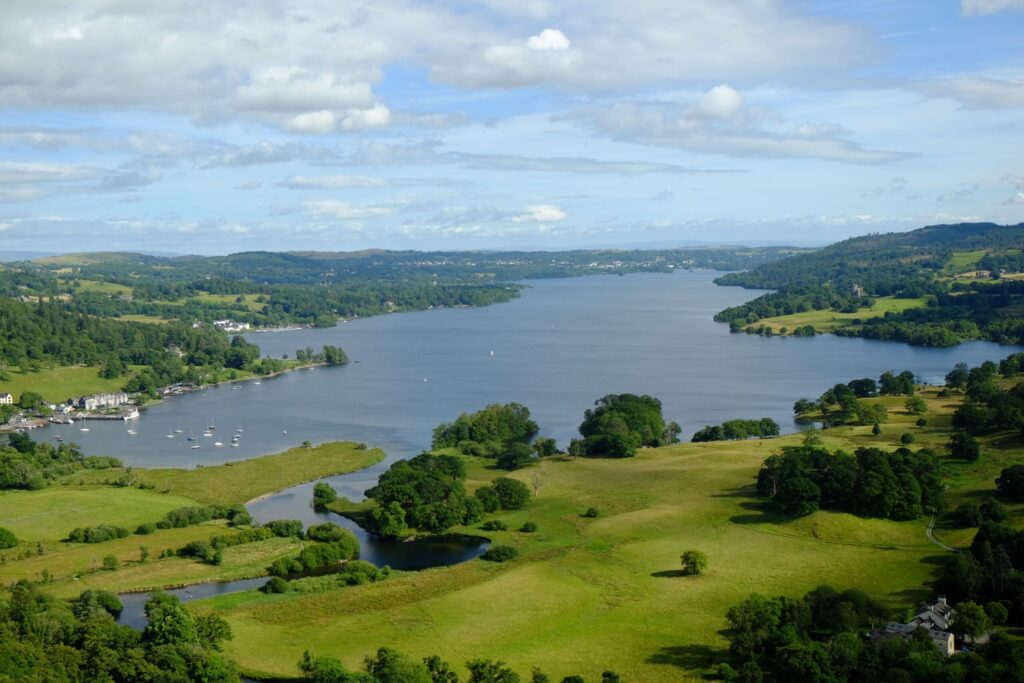 A beautiful view of Windermere lake surrounded by trees and green grass under the clouds of the sky