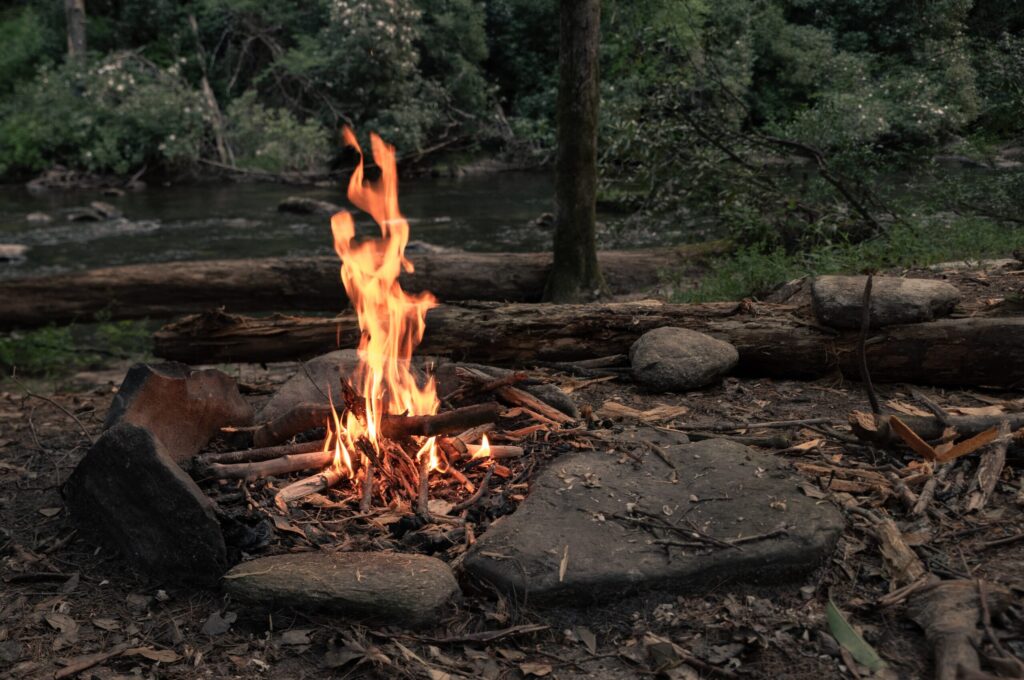 Campfire burning beside a quiet woodland river, surrounded by rocks, fallen logs and dense forest.