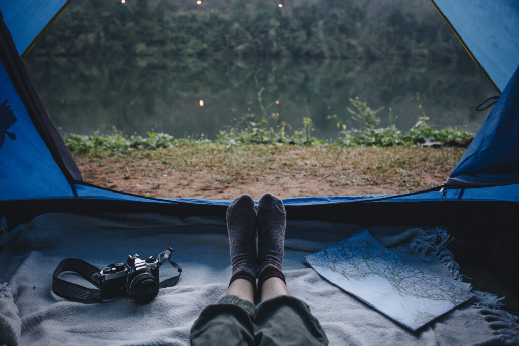 View from inside a tent by a lake, with a camper resting under a blanket beside a camera and map.