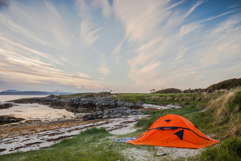 Alt tag:
Orange tent pitched beside a quiet rocky beach, with grassy dunes, calm sea and dramatic evening sky.