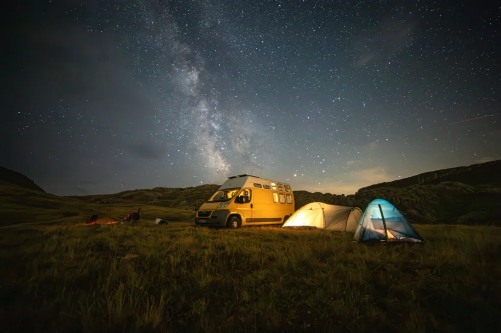 Campervan and illuminated tents beneath the Milky Way in a remote mountain campsite.