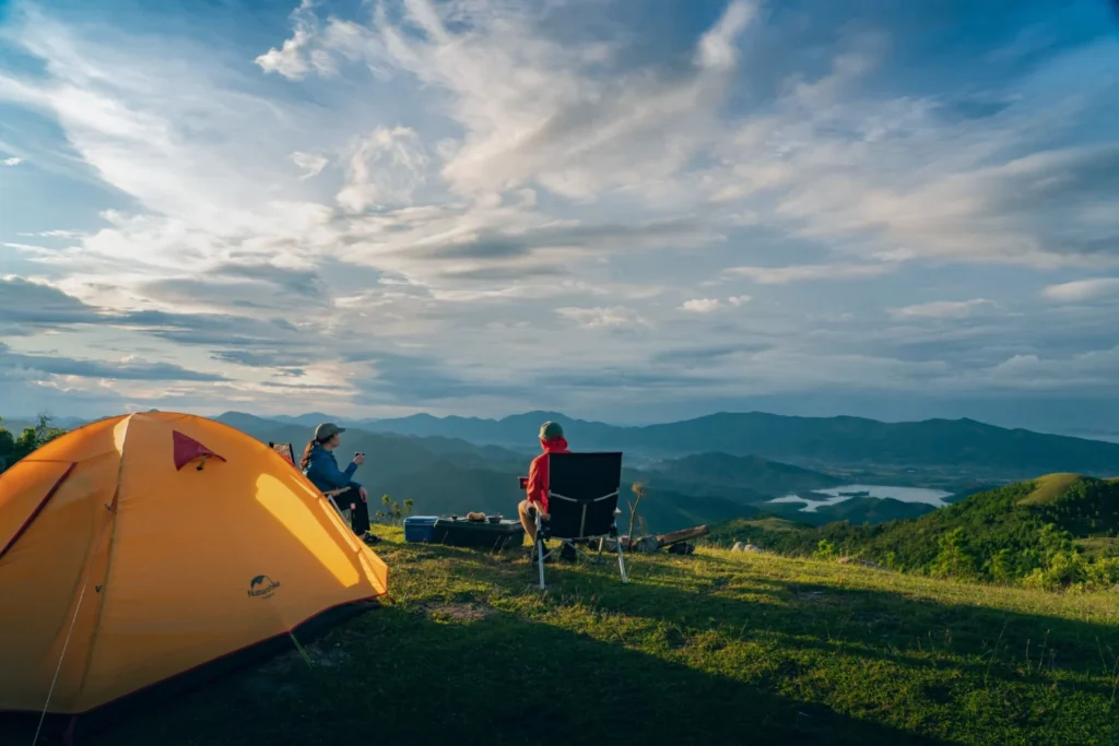 Two campers relaxing beside an orange tent overlooking mountains and a lake at sunset.
