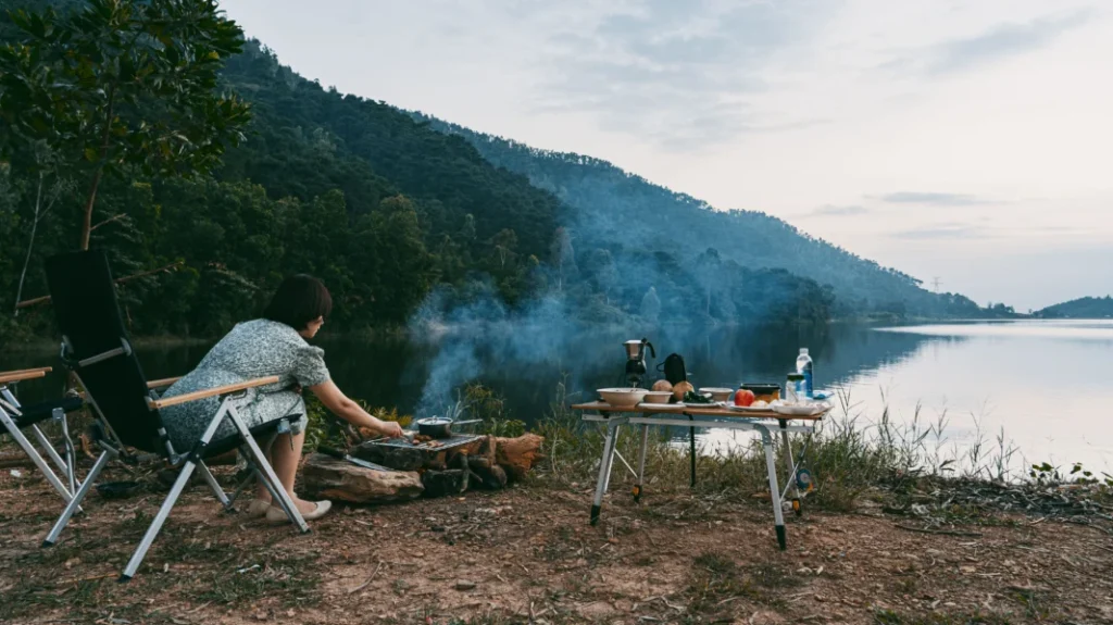 Camper cooking over a small fire beside a camping table overlooking a quiet mountain lake.